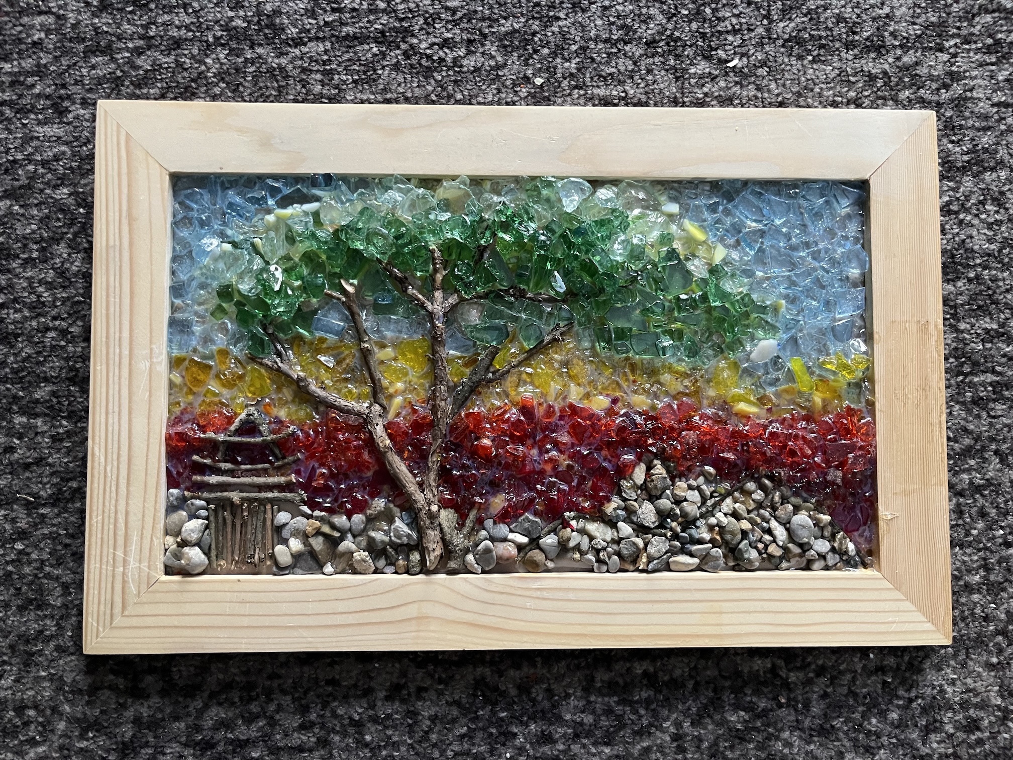 A tree with a sunset in the background made of stones, stained glass, and sticks inside a wood frame.