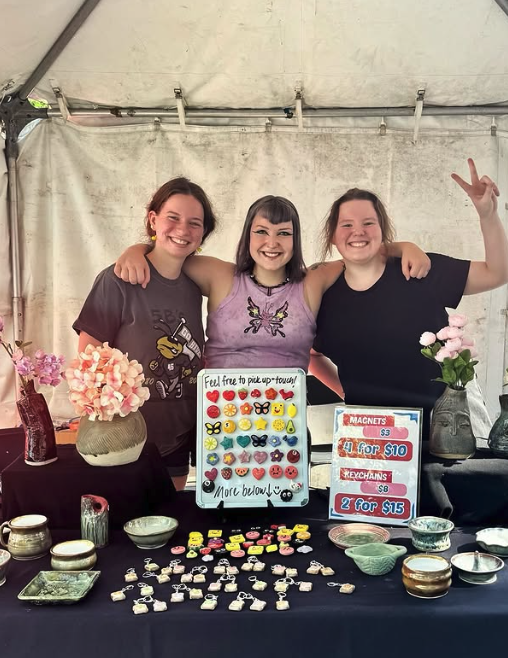 Three YSU students posing and smiling in their tent. Their art work is on a table in front of them.